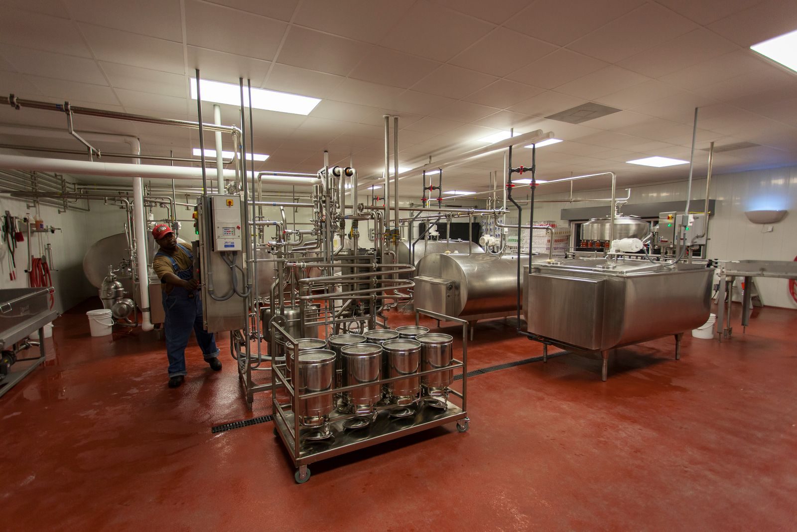 Un opérateur en salopette travaille près d'une armoire de commande dans la salle de production d'une laiterie, entre des cuves et des tuyauteries en inox et un chariot de bidons métalliques.