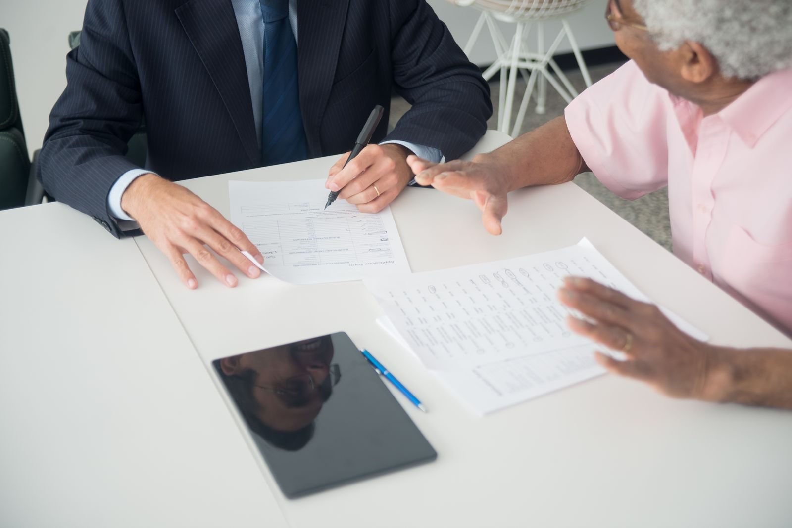 Un conseiller en costume, assis à un bureau, tient un stylo au-dessus d'un formulaire tourné vers un client âgé, qui tient d'autres documents et commente d'un geste de la main.