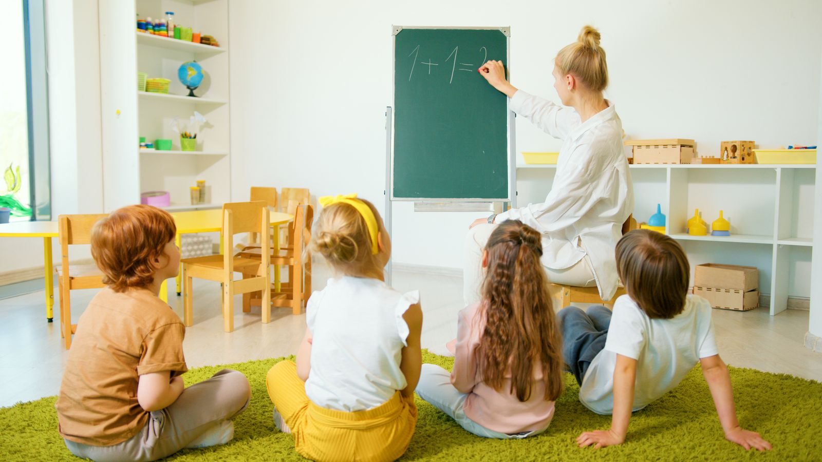 Une enseignante écrit l'opération 1 + 1 = 2 à la craie sur un tableau monté sur chevalet, devant quatre jeunes enfants assis de dos sur un tapis vert.