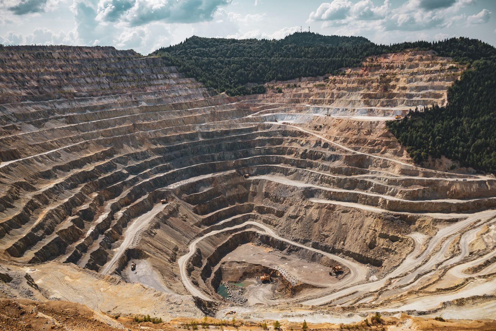 Blick von oben auf einen weitläufigen Tagebau mit terrassenförmigen Abbaustufen, umgeben von Wald, mit Baumaschinen auf der Sohle der Grube.