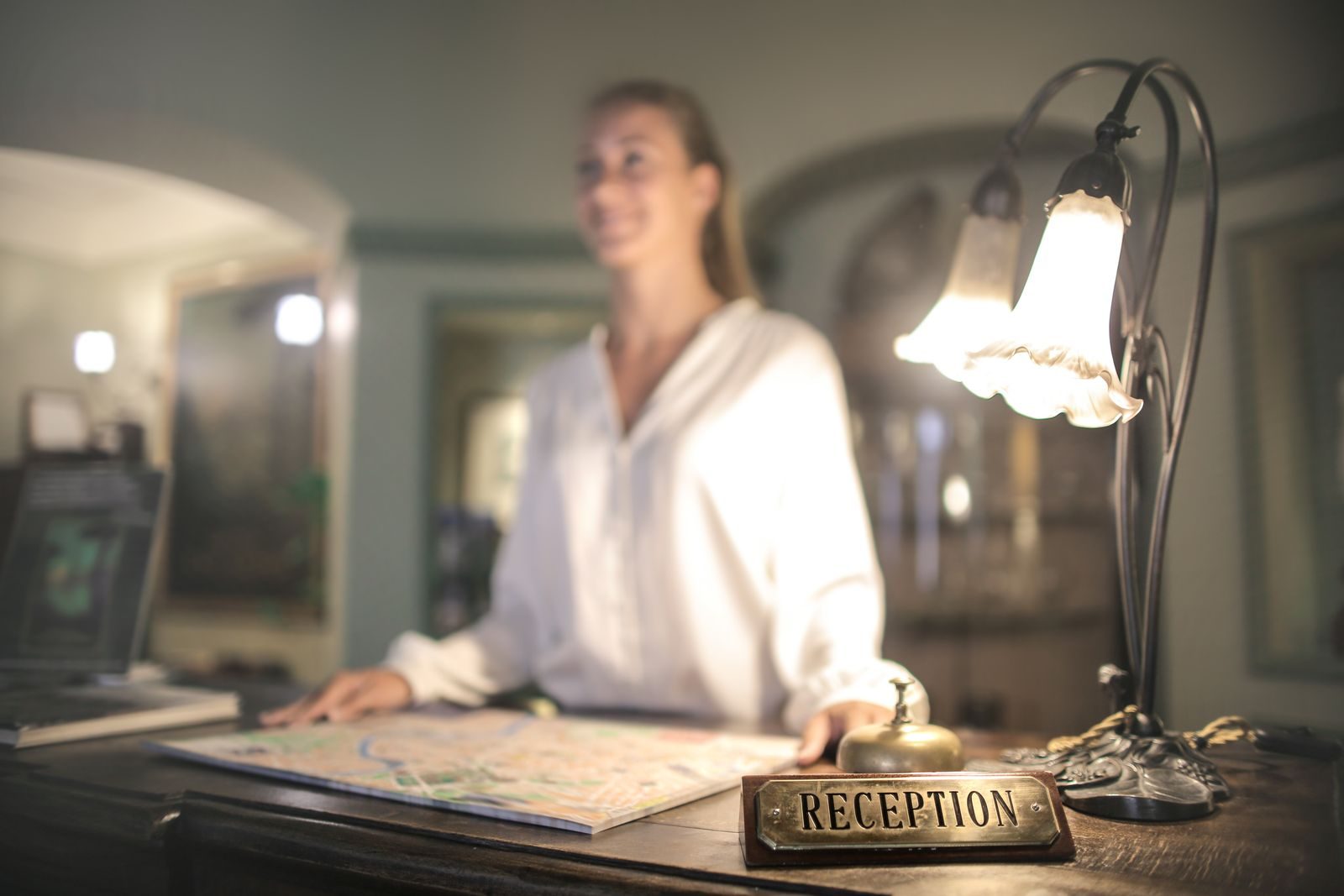 Der Holztresen einer kleinen Hotelrezeption mit Messingschild «Reception», Tischglocke und aufgefaltetem Stadtplan; dahinter eine lächelnde Rezeptionistin.