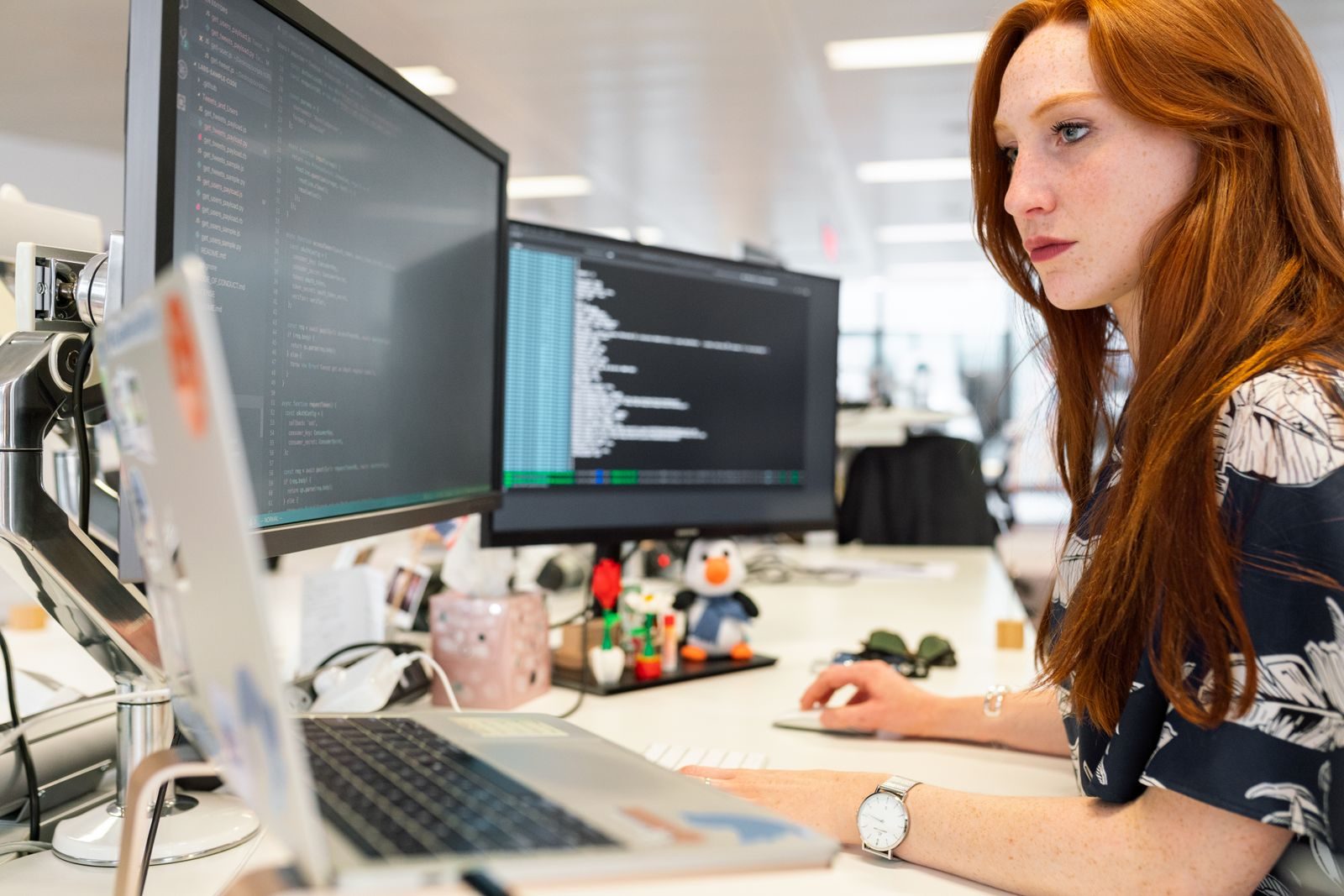 Une développeuse travaille devant deux écrans affichant du code et un terminal, dans un bureau ouvert lumineux.