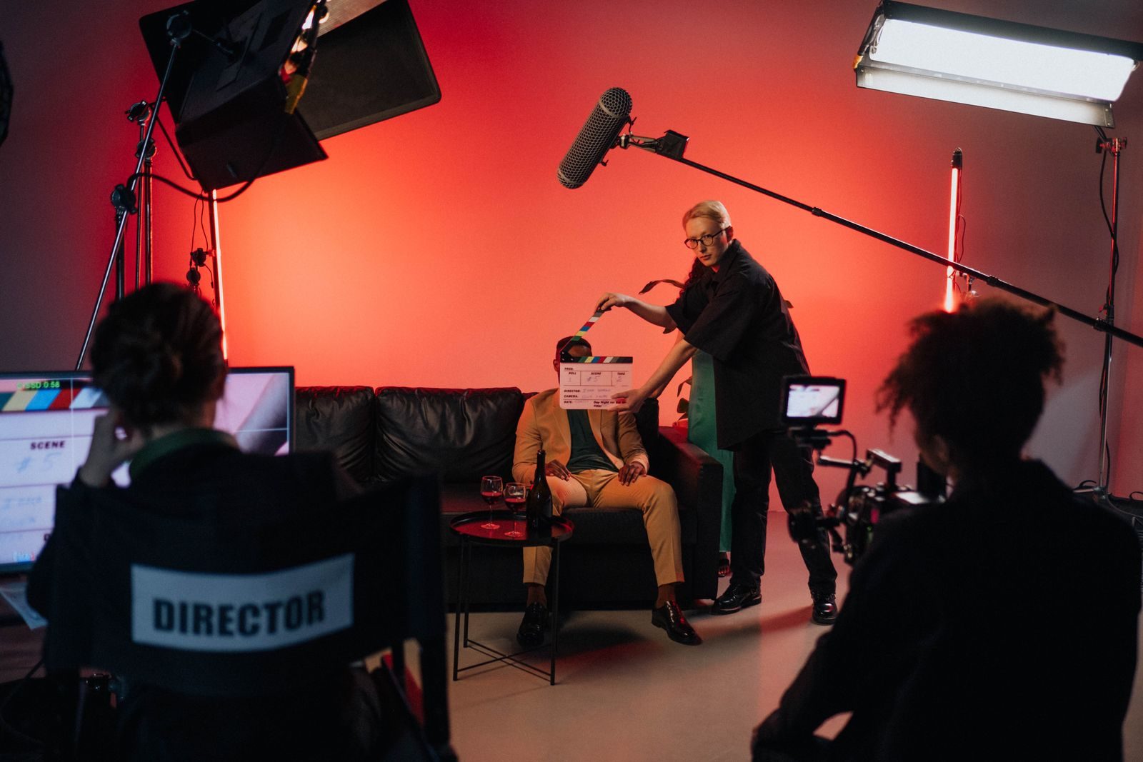 Auf einem rot ausgeleuchteten Filmset hält ein Crewmitglied die Klappe vor einen Schauspieler auf einem Sofa, während eine Kamera und ein Kontrollmonitor die Szene verfolgen.