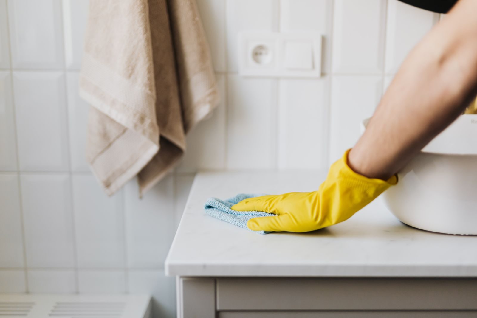 Une main gantée de jaune essuie un plan de travail blanc avec une lingette microfibre dans une salle de bains carrelée.