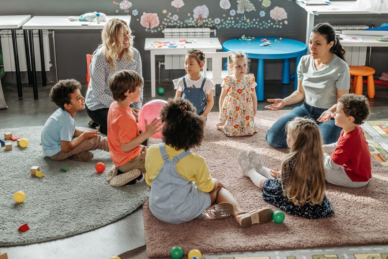 Deux éducatrices de la petite enfance assises par terre avec un groupe de jeunes enfants réunis en cercle sur un tapis, dans une salle de crèche colorée où sont éparpillés des cubes en bois et des ballons.