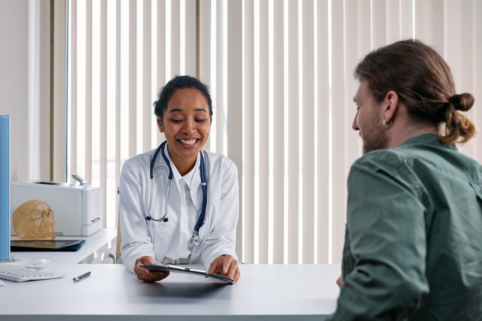 Eine Ärztin im weissen Kittel mit Stethoskop zeigt einem ihr gegenübersitzenden Patienten im Sprechzimmer ein Tablet.