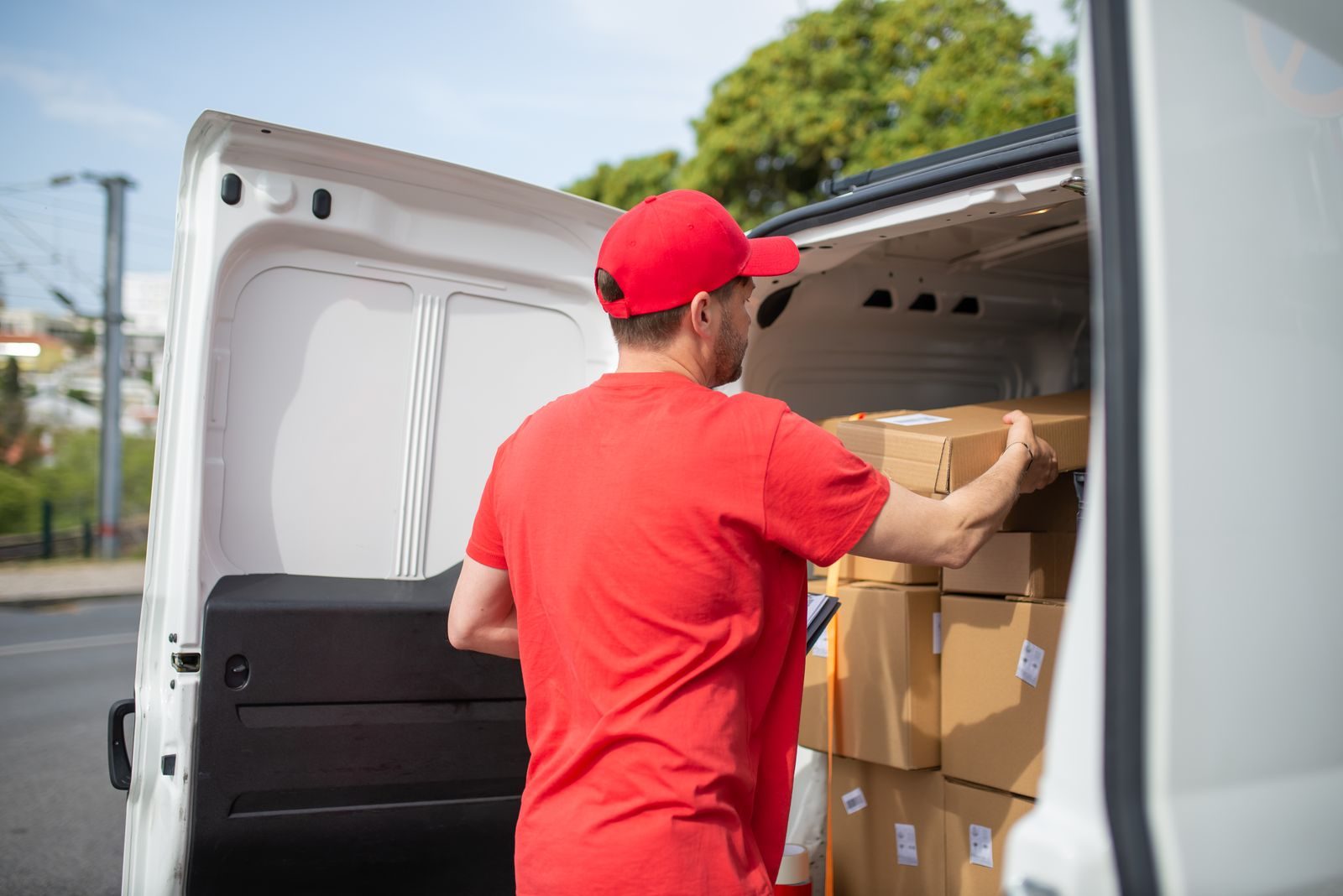 Un livreur en t-shirt et casquette rouges, vu de dos, charge un carton étiqueté dans un fourgon blanc dont les portes arrière sont ouvertes.